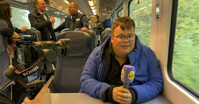 Charlie sits on a train looking relaxed and holding a microphone while a camera films him.
