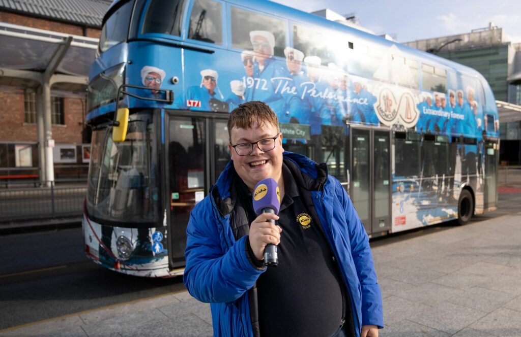 Charlie stands in front of a London bus holding a KIDS-branded microphone and smiling