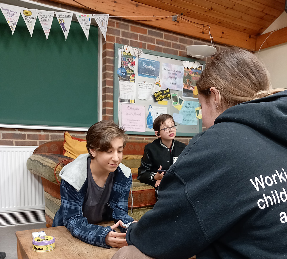 Two young carers play cards while another sits on a sofa holding a phone