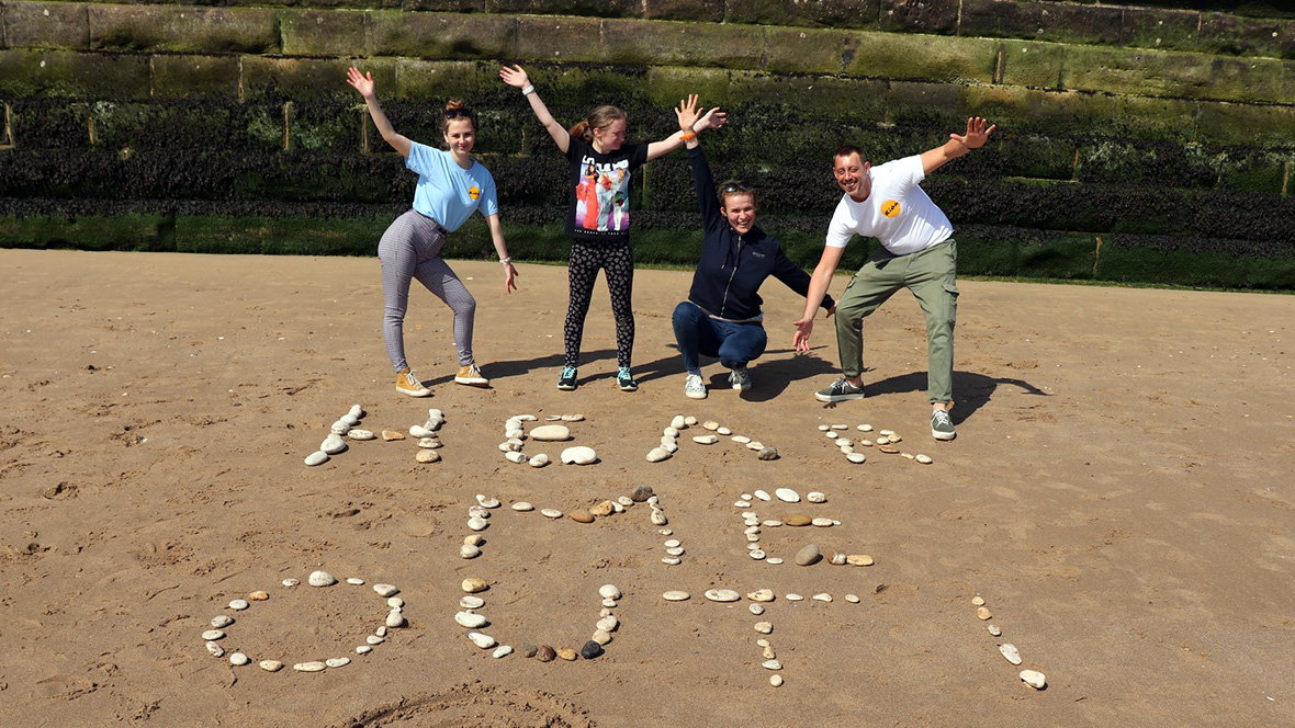 Two young people and two adults stand waving on a beach with 'Hear Me Out' written in pebbles in front of them.